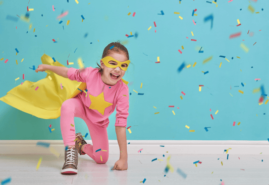 A young girl in a pink super suit with a yellow star on her chest and a yellow cape on her back. She is on one knee with a fist on the ground, a classic superhero pose. There is a baby blue background and confetti falling around her.