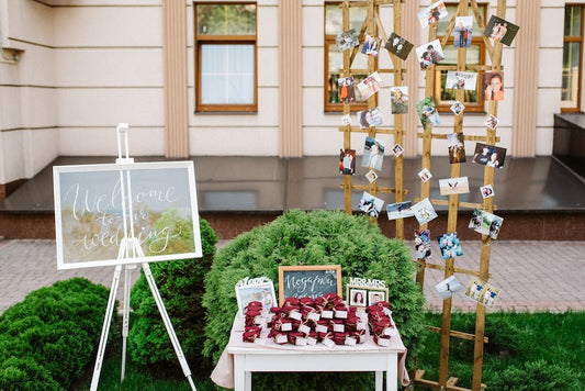 A small white table set up in front of green bushes. There are little gifts wrapped in red cloth. There is a white sign on a white easel that says "Welcome to our wedding". There is also a wooden backdrop with pictures of the bride and groom attached.