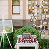A small white table set up in front of green bushes. There are little gifts wrapped in red cloth. There is a white sign on a white easel that says 
